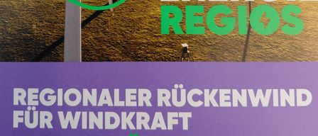 Ein Bild zeigt zwei Windräder auf einem grünen Feld mit dem Text 'Unsere Energie Regios' und 'Regionaler Ruckenwind für Windkraft' in Deutsch.