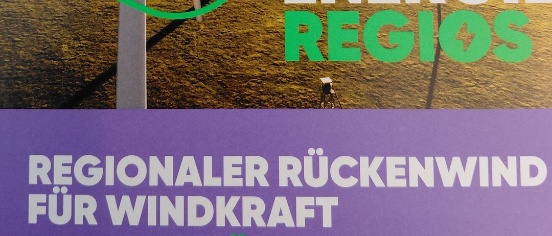 Ein Werbeposter zeigt ein Feld mit zwei Windturbinen. Der Plakattext lautet 'Unsere Energie Regios' und 'Regionaler Ruckenwind fur Windkraft.' Eine grüne Linie führt von den Turbinen zum Text.