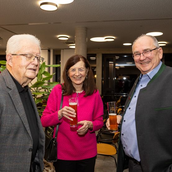Drei Personen stehen in einem Innenraum zusammen und lächeln für ein Foto. Die Frau in der Mitte hält ein Glas Bier. Der Mann rechts trägt eine Brille und eine grüne Schleife, während der Mann links einen grauen Mantel trägt. Hinter ihnen steht eine Pflanze und Stühle.