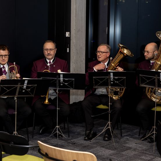 Vier Männer in formeller Kleidung sitzen in einem Raum und spielen Blechblasinstrumente. Zwei halten Trompeten, einer hält eine Tuba und der vierte hält ein Waldhorn.
