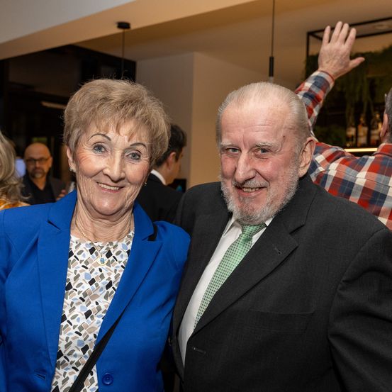 Ein älteres Paar lächelt für ein Foto bei einer gesellschaftlichen Veranstaltung. Die Frau trägt einen blauen Blazer und der Mann ein schwarzes Kostüm mit grüner Krawatte. Andere Menschen sind im Hintergrund zu sehen.