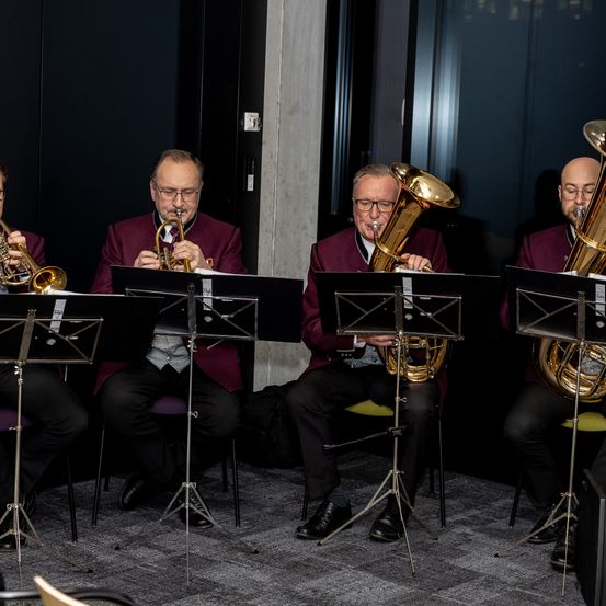 Vier Musiker sitzen auf Stühlen und spielen Trompeten und Tubas. Sie befinden sich in einem Raum mit dunklem Hintergrund. Alle tragen formelle Kleidung und Brillen.