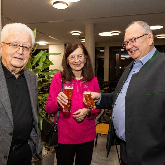 Drei Personen posieren für ein Foto in einem Raum und halten Gläser mit Bier in den Händen. Die Frau in der Mitte trägt einen rosa Pullover und eine Halskette. Beide Männer lächeln. Im Hintergrund befinden sich eine Pflanze und eine Säule.