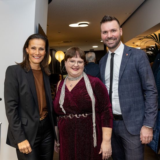 Drei Personen, zwei Frauen und ein Mann, stehen lächelnd zusammen in einem Raum. Die Frau auf der linken Seite trägt einen schwarzen Blazer. Die mittlere Frau hat ein burgunderrotes Kleid, eine Brille und einen silbernen Schal. Der Mann auf der rechten Seite trägt einen marineblauen Anzug und eine Krawatte.