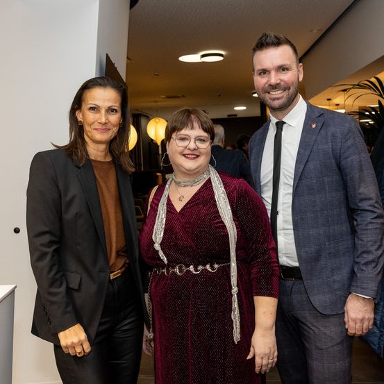 Drei Menschen stehen für ein Foto zusammen, zwei Frauen und ein Mann. Die Frauen tragen Brillen und Halsketten. Sie sind in formeller Kleidung gekleidet.