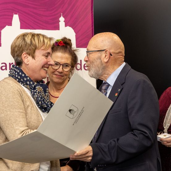 Drei Personen versammeln sich in einem Raum; ein Mann und zwei Frauen, eine hält eine Urkunde. Sie wirken fröhlich, mit Lächeln auf ihren Gesichtern. Hinter ihnen zeigt ein Hintergrund ein rosafarbenes Banner mit einem Schlossdesign.
