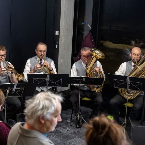 Eine Gruppe von Musikern, alle mit Brille, spielt Blechblasinstrumente in einem Raum mit Glasfenster. Eine Person beobachtet sie.