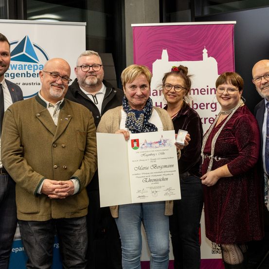 Eine Gruppe von Menschen, darunter eine Frau mit einem Zertifikat, steht gemeinsam vor einem Banner, möglicherweise bei einer Preisverleihung.
