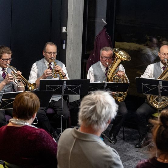 Ein Blechbläserensemble, bestehend aus Trompeten und Tubas, spielt in einem Raum mit schwarzen Wänden. Die Musiker sitzen auf Stühlen, und das Publikum beobachtet sie aufmerksam.