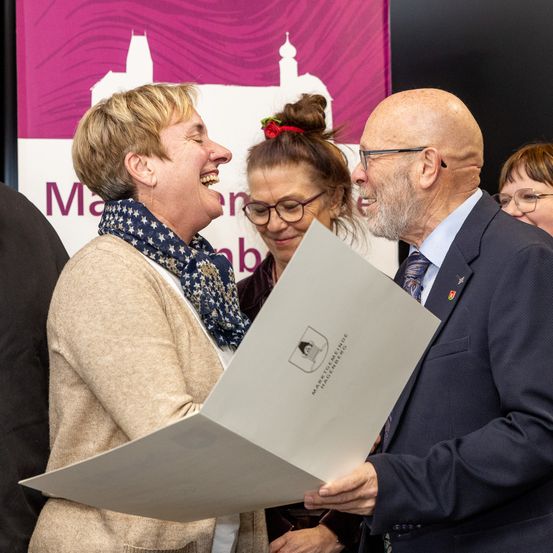 Eine Gruppe von Menschen steht vor einem Banner. Eine Frau hält ein Papier und lacht. Ein Mann mit Bart und Brille lächelt und schaut auf das Papier.