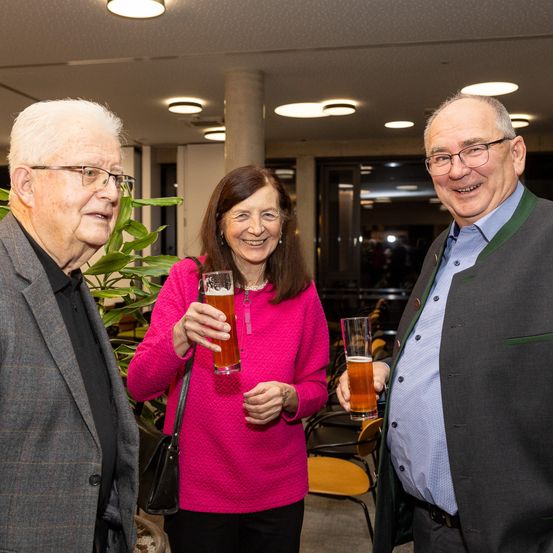 Drei Personen stehen eng beieinander und halten Gläser mit Bier. Der Mann links trägt eine Brille und einen Anzug. Die Frau in der Mitte trägt einen rosa Pullover und trägt eine schwarze Tasche. Der Mann rechts trägt eine grüne und graue Weste.