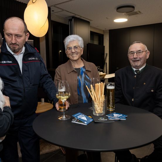 Vier Menschen versammeln sich um einen Tisch, zwei Männer und zwei Frauen. Die Männer tragen Jacken mit Logos, und die Frauen tragen Brillen und Jacken. Gläser und Strohhalme stehen auf dem Tisch.