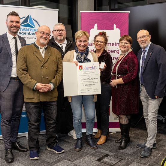Eine Gruppe von Menschen steht lächelnd bei einer Veranstaltung, hält ein Zertifikat und eine Medaille. Die Frau in der Mitte hält das Zertifikat und die Medaille, während andere hinter ihr stehen, lächeln und in die Kamera schauen.