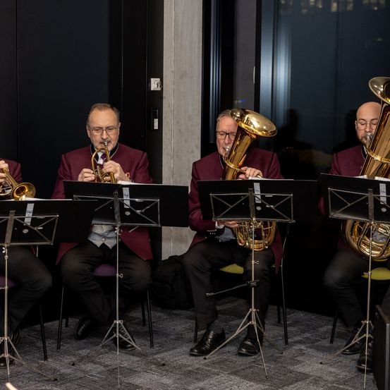 Vier Musiker sitzen und spielen ihre Instrumente. Einer ist auf der Trompete, zwei auf Tuben und einer auf einem Posaune. Sie tragen aufeinander abgestimmte burgunderrote Jacken und konzentrieren sich auf ihre Notenblätter.