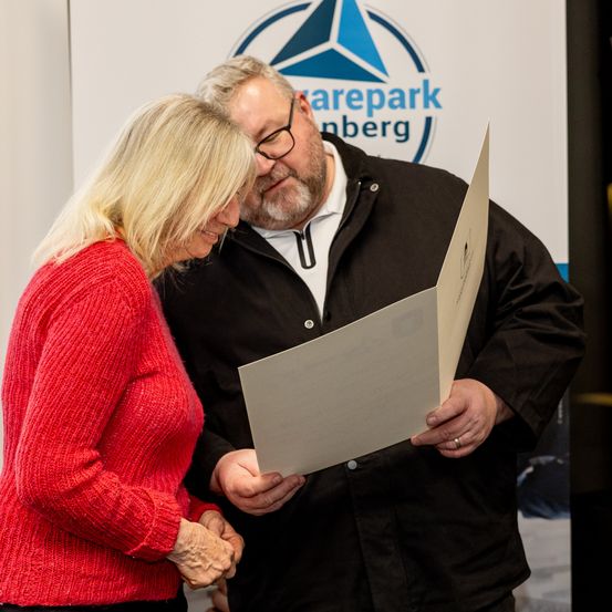 Ein Mann und eine Frau stehen nah beieinander und schauen sich ein Dokument an, das der Mann hält. Sie lächeln. Dahinter befindet sich ein weißer Banner mit einem Logo und Text.