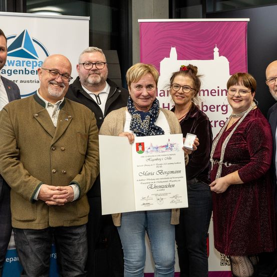 Eine Gruppe von Menschen lächelt und posiert für ein Foto mit einem Zertifikat. Eine Frau in der Mitte hält das Zertifikat mit beiden Händen. Hinter ihnen befindet sich ein rosafarbener Banner mit Text.