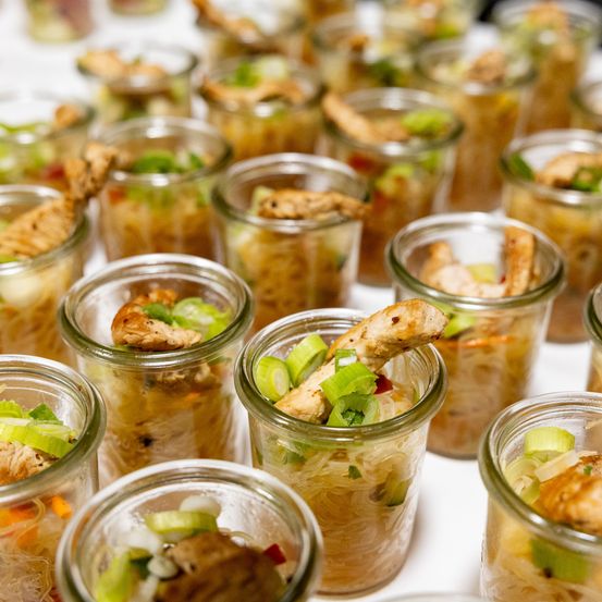 Eine Nahaufnahme von mehreren kleinen Glasgläsern, gefüllt mit Hühnchen- und Gemüsenudelsuppe. Die Suppe enthält gehacktes Hühnchen, Frühlingszwiebeln und andere Gemüse.