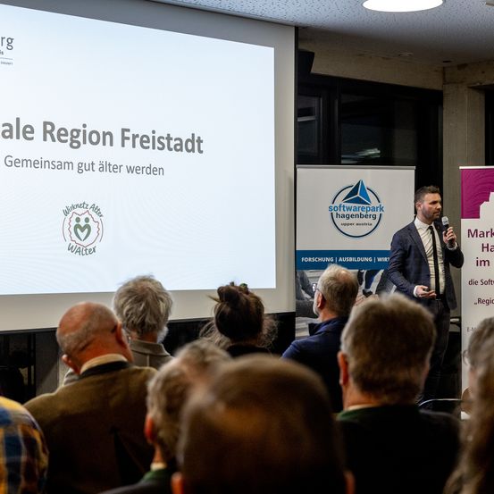 Ein Mann spricht in ein Mikrofon vor einer Versammlung. Hinter ihm zeigt eine Leinwand eine Präsentation mit dem Titel 'sale Region Freistadt'. Der Mann trägt einen Anzug und eine Krawatte. Das Publikum besteht aus verschiedenen Menschen in Freizeitkleidung. Ein Banner mit dem Text 'Softwarepark Hagenberg' ist ebenfalls zu sehen.