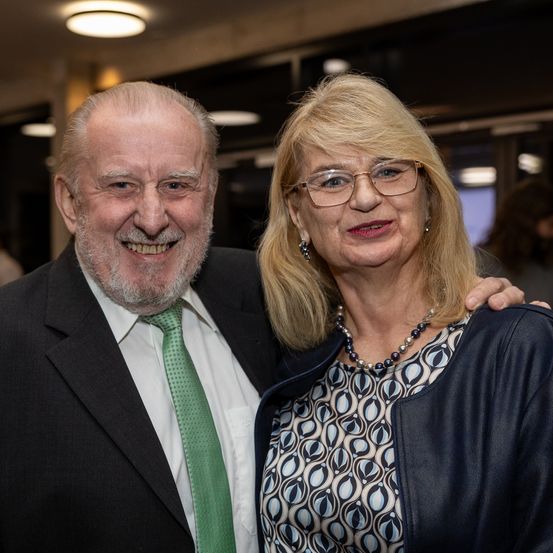Ein älterer Mann und eine Frau lächeln für ein Foto. Der Mann trägt eine grüne Krawatte und die Frau eine Brille und eine Halskette. Sie stehen eng beieinander, wobei eine weitere Person im Hintergrund zu sehen ist.