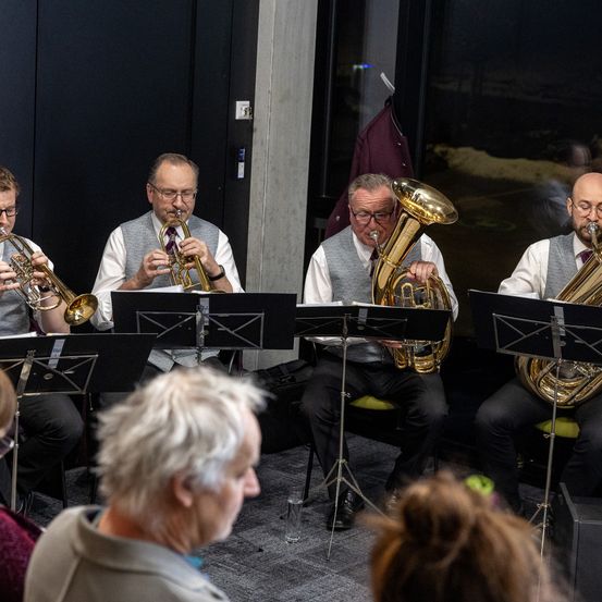 Vier Musiker, zwei Trompeter und zwei Tubaspieler, spielen auf der Bühne vor einem Publikum. Sie sind in formeller Kleidung gekleidet und von Notenständern umgeben.