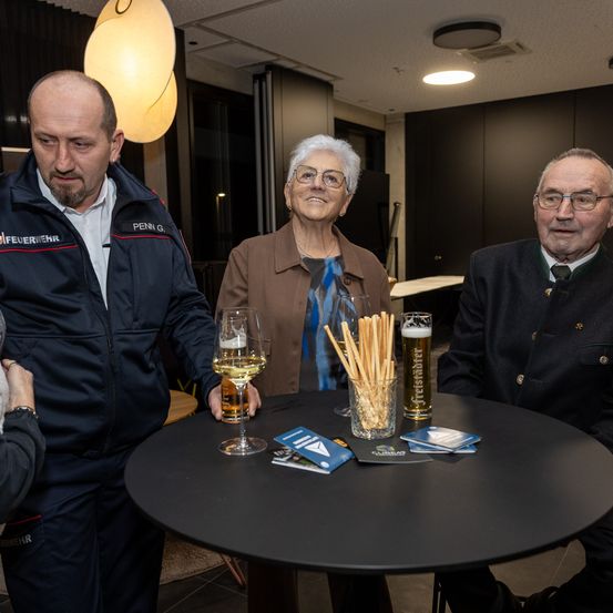 Drei Personen stehen um einen Tisch mit Biergläsern. Der Mann links trägt eine Jacke mit Logo. Die Frau in der Mitte lächelt und der Mann rechts trägt eine Brille.
