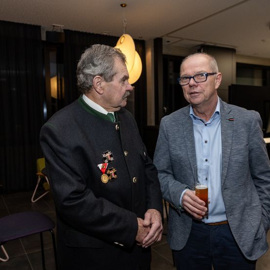 Zwei Männer stehen nah beieinander, einer mit Medaillen und einem Getränk, der andere in einem Anzug, unterhalten sich drinnen mit Stühlen und einer Lampe im Hintergrund.