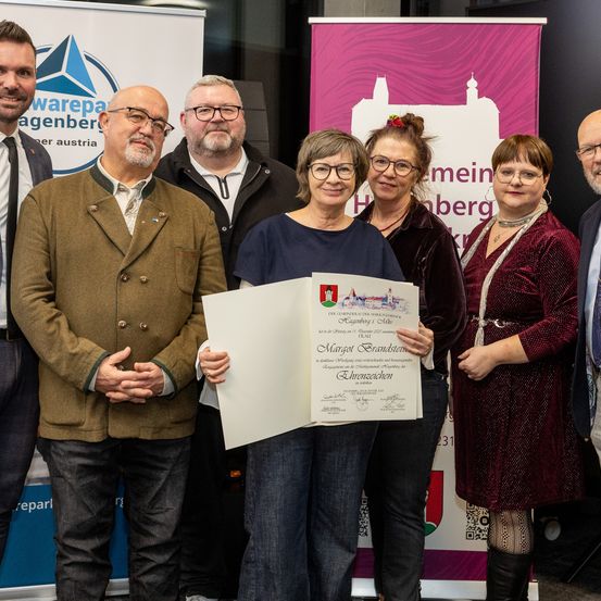 Eine Gruppe von Menschen steht vor einem Banner und hält eine Urkunde. Die Frau in der Mitte hält sie mit beiden Händen und lächelt. Hinter ihr stehen zwei Frauen, eine mit einer Halskette. Sie werden von drei Männern umgeben, zwei in Anzügen. Das Banner zeigt ein Logo und Text.