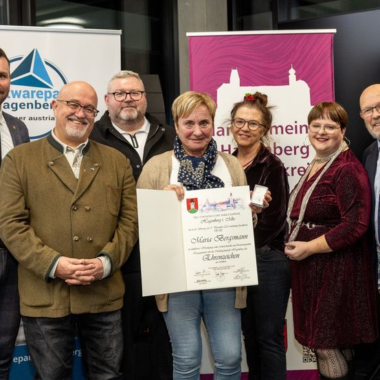 Eine Gruppe von Menschen, darunter zwei Frauen, eine mit einem Zertifikat in der Hand, steht vor einem Banner mit den Worten 'mein berg' für ein Foto.