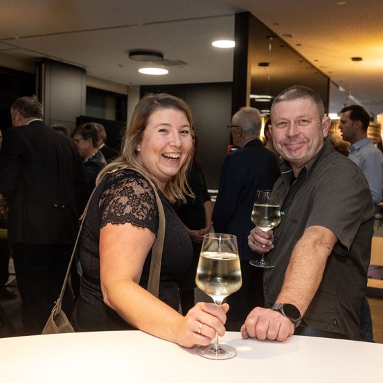 Eine lächelnde Frau und ein Mann, beide mit Weinglas in der Hand, stehen vor einem Tisch, während andere Menschen im Hintergrund sind, einige verschwommen.