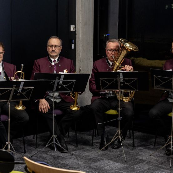 Vier Musiker in Purpuruniformen sitzen und spielen Instrumente. Zwei spielen Trompeten und zwei spielen Tuben. Sie sind vor Notenständern in einem schwach beleuchteten Raum positioniert.