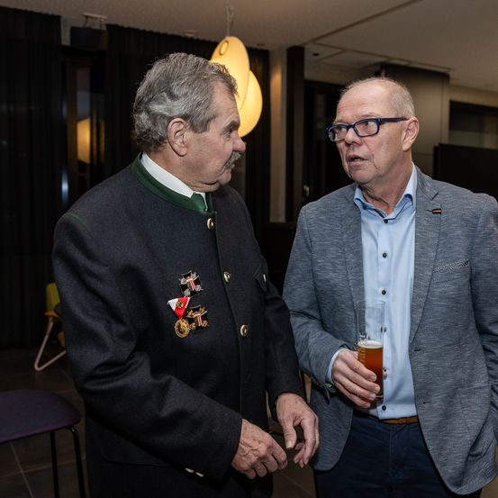 Zwei Männer stehen in einer Innenumgebung, einer trägt eine mit Medaillen verzierte Jacke und der andere hält ein Glas Bier. Der Mann auf der rechten Seite trägt eine Brille und eine graue Anzugjacke.