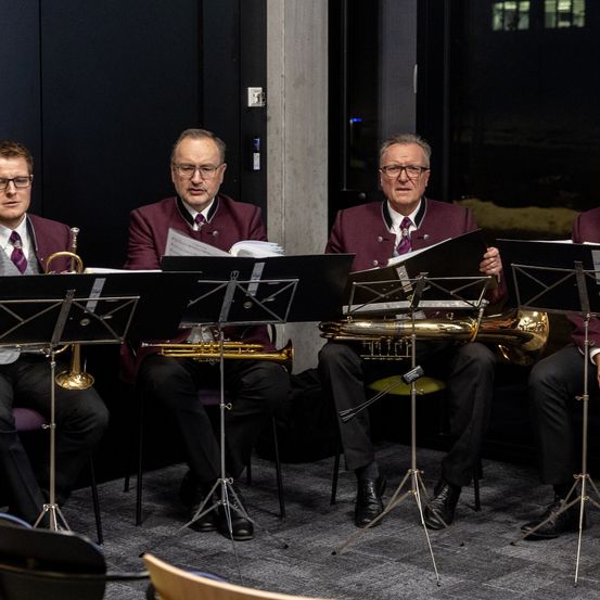Vier Musiker sitzen auf Stühlen in einem schwach beleuchteten Raum und halten jeweils ein Tenorhorn und lesen Musik. Sie sind in formeller Kleidung gekleidet, was auf eine ernste Aufführung hindeutet.