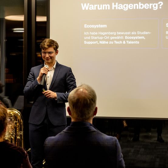 Ein Mann hält eine Rede vor einem Publikum und hält ein Mikrofon in der Hand. Hinter ihm ist eine Präsentationsfolie sichtbar, die eine Frage zu Hagenberg stellt.