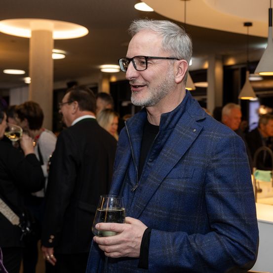Ein Mann in einem blauen Anzug und mit Brille lächelt und hält ein Glas Wein bei einer Veranstaltung. Mehrere Personen stehen hinter ihm.