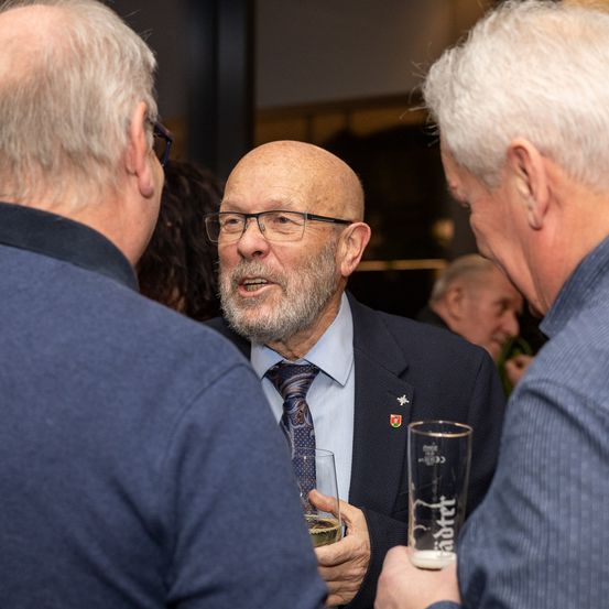 Ein älterer Mann mit Brille und Krawatte führt ein Gespräch mit zwei anderen Männern bei einer gesellschaftlichen Veranstaltung. Er hält ein Glas Wein in der Hand.