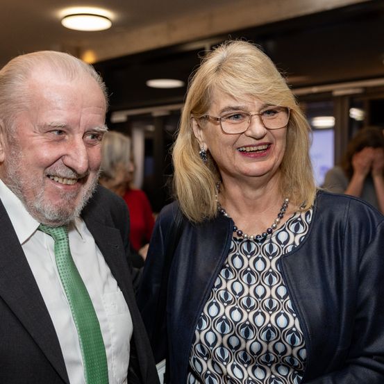 Ein älterer Mann und eine Frau lächeln und posieren für ein Foto. Der Mann trägt eine grüne Krawatte und die Frau eine Brille und eine Perlenkette. Sie befinden sich in einem Raum mit beleuchteten Decken.