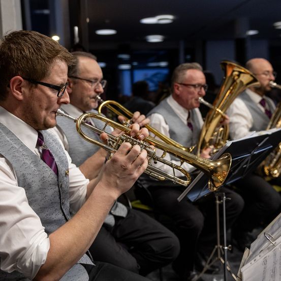 Vier Männer spielen Blechblasinstrumente in einem schwach beleuchteten Raum. Ein Mann spielt Trompete, ein anderer Posaune und zwei weitere spielen Tuba.