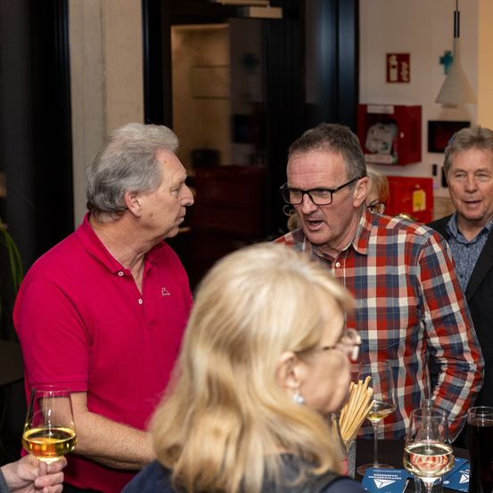 Eine Gruppe älterer Personen unterhält sich in einem Innenraum. Ein Mann trägt ein rotes Poloshirt, ein anderer ein kariertes Hemd und ein dritter einen dunklen Anzug. Brillen, Weingläser und ein roter Feuerlöscher sind sichtbar.