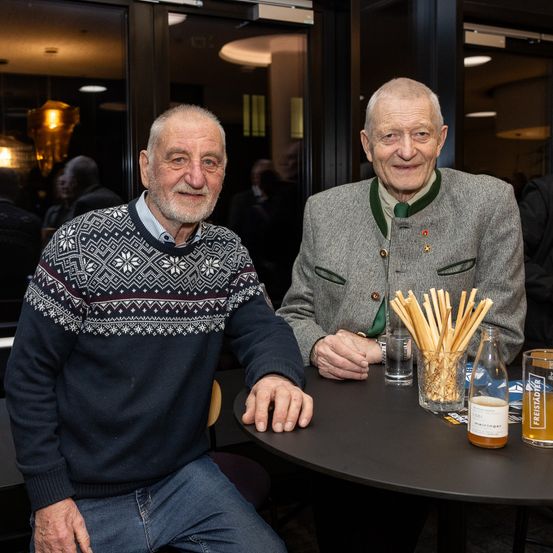 Zwei ältere Männer sitzen an einem Tisch, lächeln und posieren für ein Foto. Sie sind von Gläsern und einer Flasche Bier umgeben. Im Hintergrund sitzen Leute an einem Tisch, und eine Lampe hängt von der Decke.
