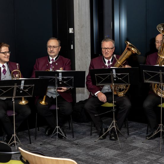 Vier Musiker sitzen und spielen Blechblasinstrumente, wahrscheinlich in einer Probe. Sie tragen passende Uniformen. Jeder hat einen Notenständer vor sich. Der Hintergrund ist dunkel.