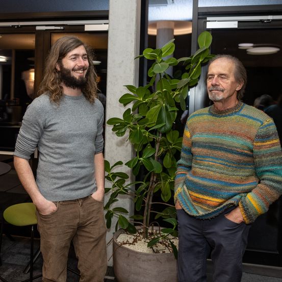 Zwei Männer stehen neben einer großen Topfpflanze in einem modernen Gebäude. Ein Mann hat lange Haare und einen Bart, während der andere einen bunten Pullover trägt.