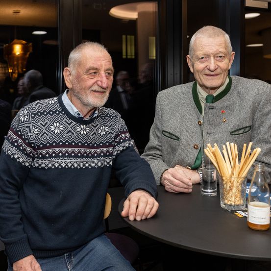 Zwei ältere Männer sitzen an einem Tisch, einer trägt einen Pullover und der andere ein traditionelles deutsches Outfit. Ein Glas Wasser und eine Glas mit Grissini sind auf dem Tisch, mit weiteren Personen im Hintergrund.