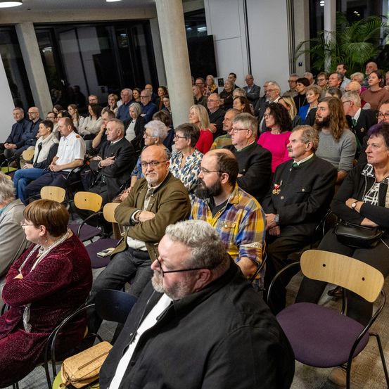 Ein volles Auditorium mit Menschen unterschiedlichen Alters und Kleidung, einige mit Brille, die einer Präsentation lauschen, mit einer Pflanze im Hintergrund.