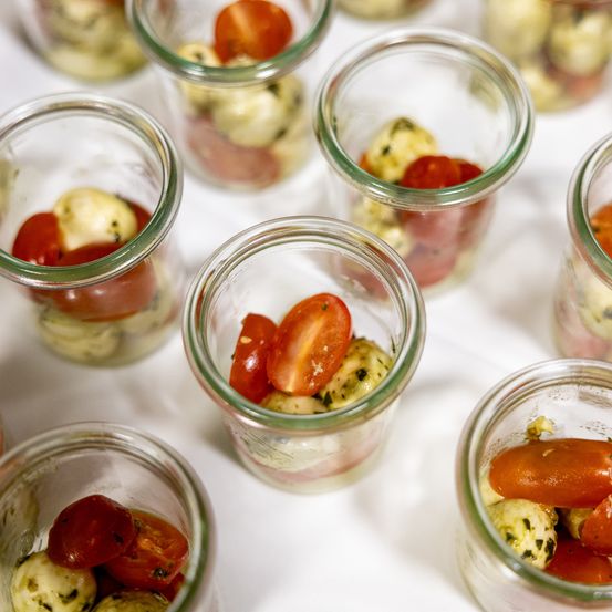 Glasbehälter gefüllt mit marinierten Mozzarellabällchen und Kirschtomaten, auf einer weißen Oberfläche präsentiert.