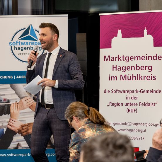 Ein Mann in einem Anzug hält eine Rede auf einer Konferenz in Hagenberg, mit einem Softwarepark-Banner im Hintergrund.