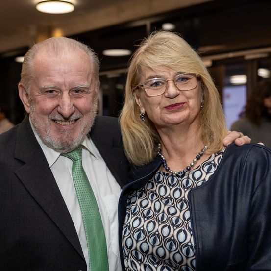 Ein älterer Mann mit grüner Krawatte und eine Frau mit Brille und Perlenkette lächeln für ein Foto.