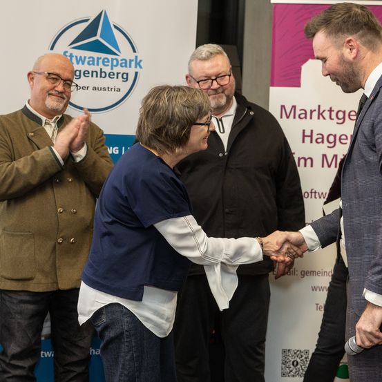 Eine Frau schüttelt einem Mann in Anzug und Krawatte die Hand. Dahinter stehen drei Männer, von denen einer applaudiert. Sie stehen vor einem Schild, das 'Softwarepark Genberg' besagt.
