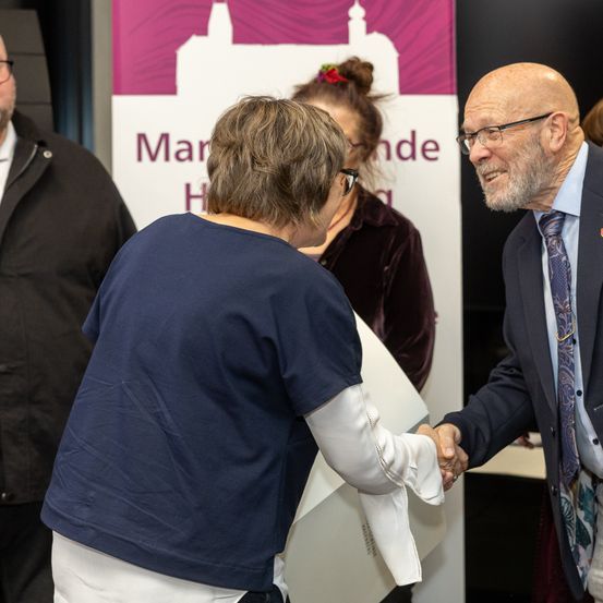 Eine Gruppe von Menschen steht bei einer Veranstaltung, wobei eine Person eine Auszeichnung an eine andere überreicht. Der Schriftzug auf dem Banner hinter ihnen lautet 'Marke H'.
