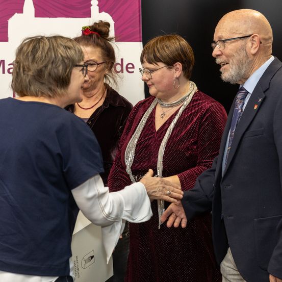 Vier Menschen stehen vor einem Banner. Eine Frau in einem roten Kleid schüttelt einem Mann im Anzug die Hand.