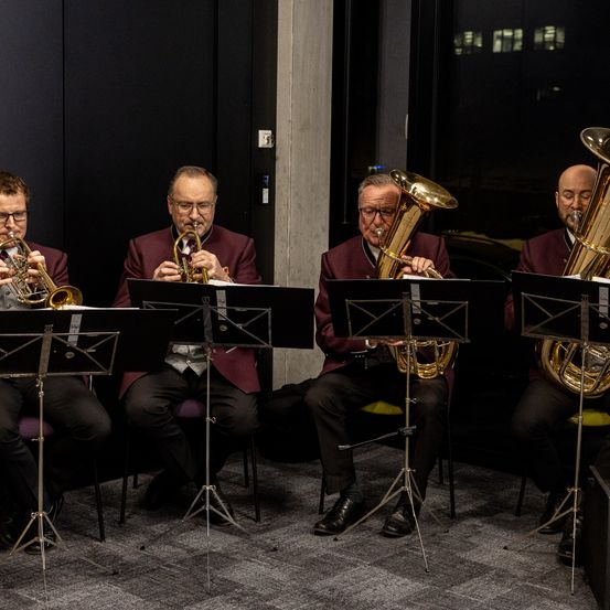 Vier Musiker sitzen und spielen Blechblasinstrumente. Einer spielt Trompete, die anderen Tuba. Sie tragen formelle Kleidung und sitzen auf Stühlen mit Notenständern vor ihnen.